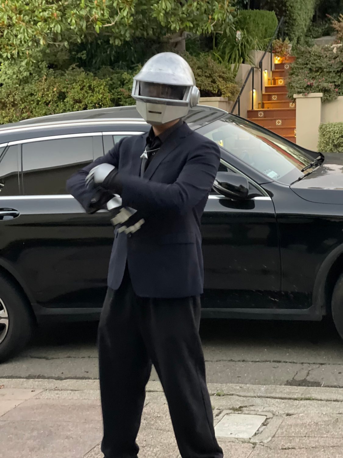Man in a dark suit wearing a silver 3D-printed Daft Punk helmet and gloves on a sidewalk