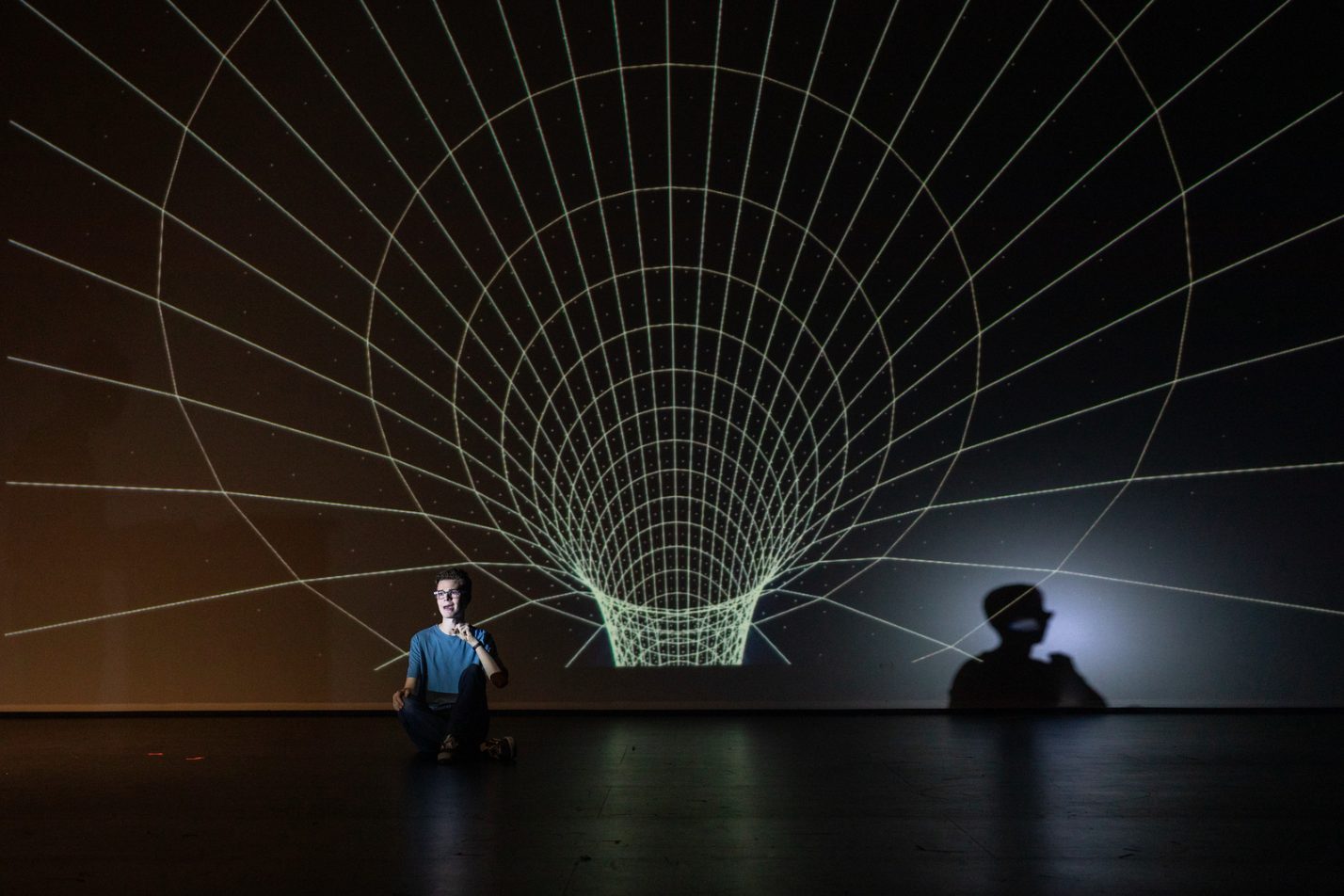 A glowing wireframe funnel projected across a theater's back wall, with a seated performer in front