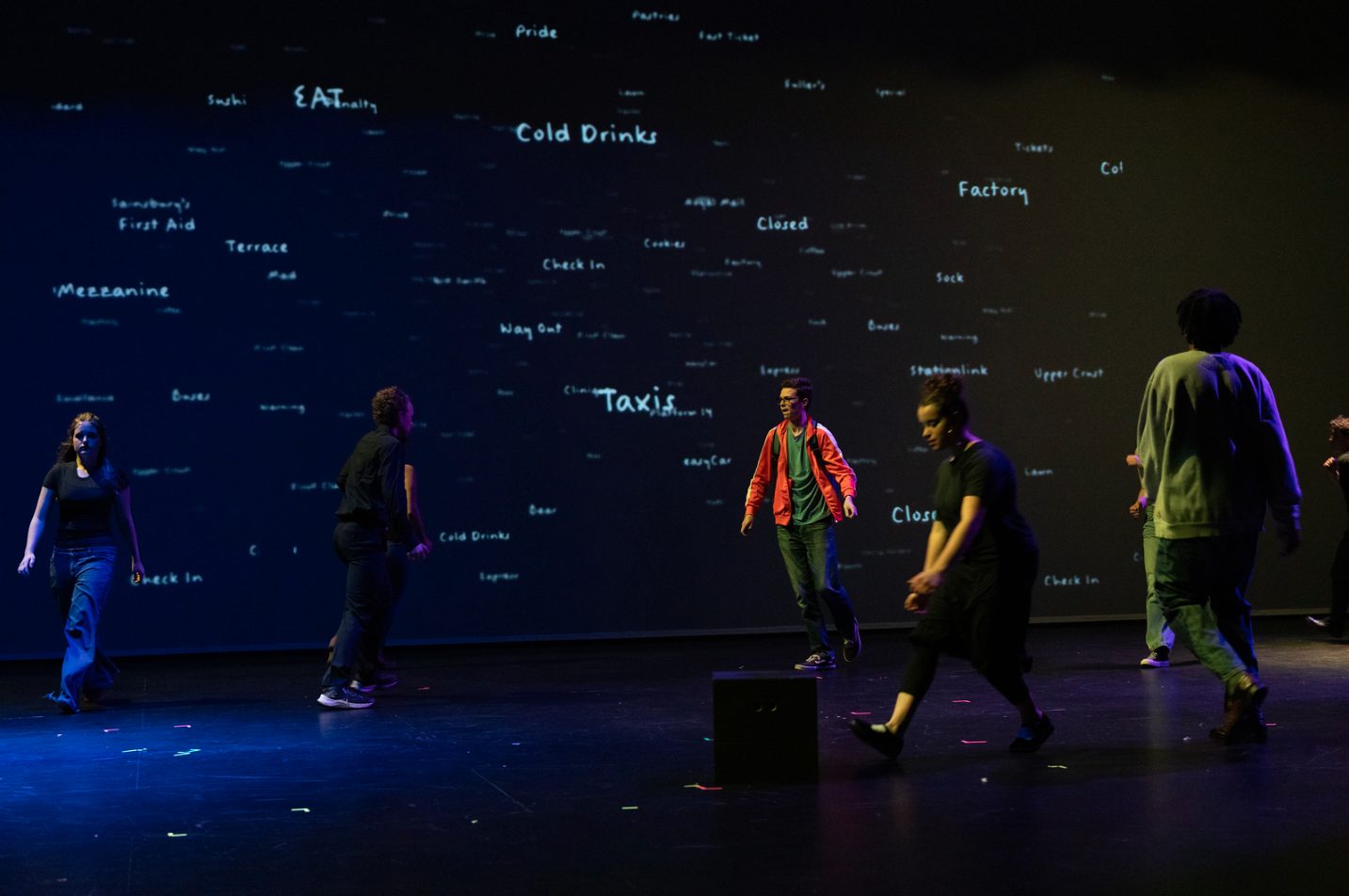 Handwritten station and shop signs scattered across a dark projected backdrop as actors cross the stage