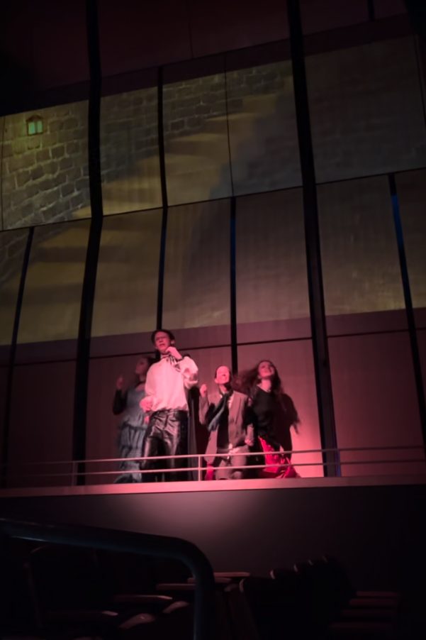 Projected stone wall with a lit window behind four performers on an upper level, lit in red